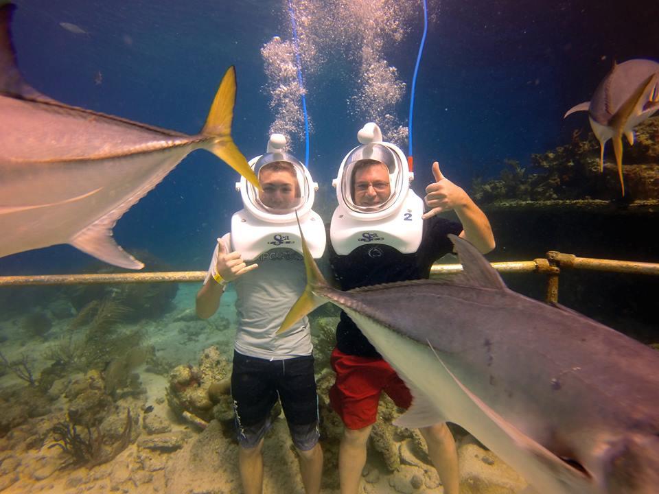 Two people underwater with helmets, surrounded by fish.
