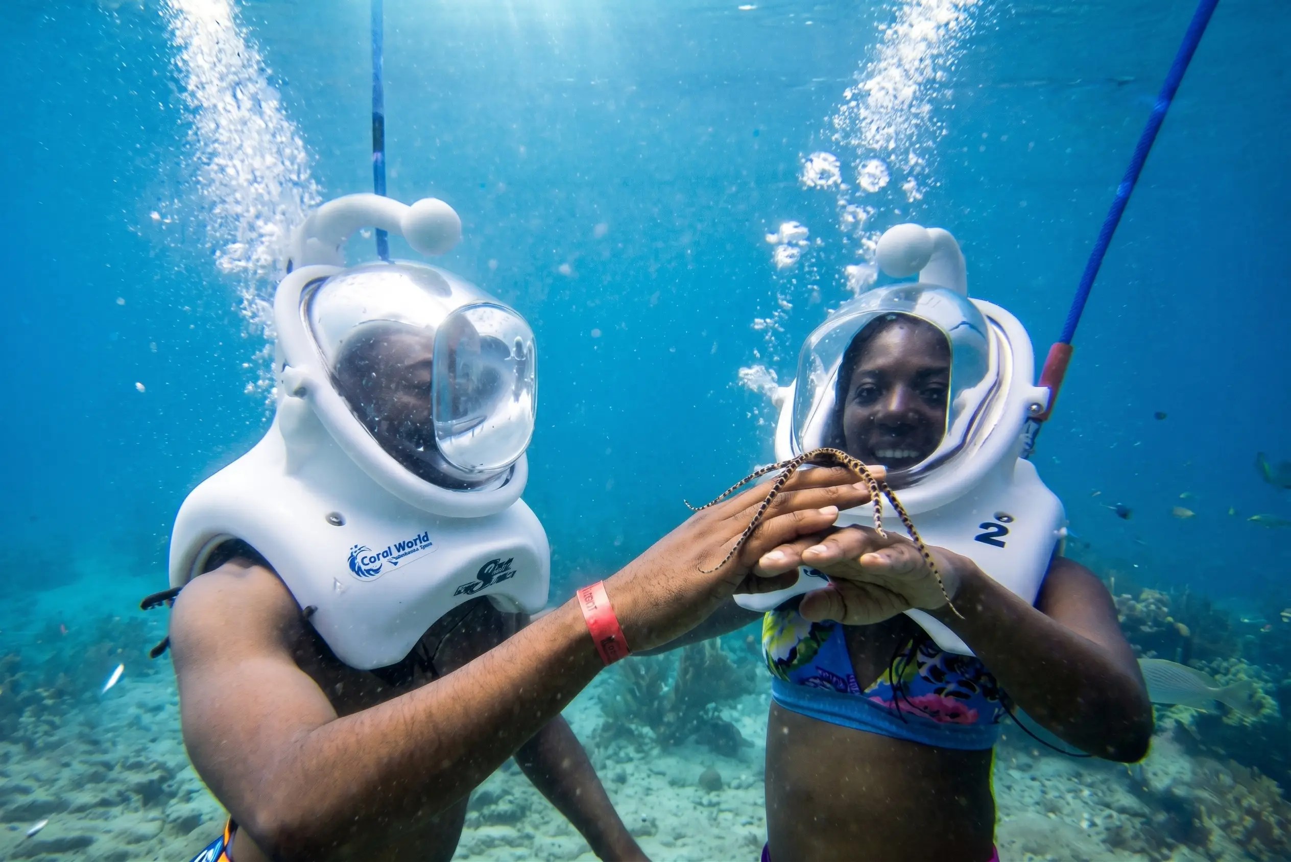 Two people underwater in dive helmets holding hands.