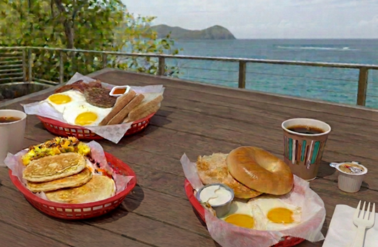 Outdoor table with breakfast plates of pancakes, bagels, and eggs near the ocean.